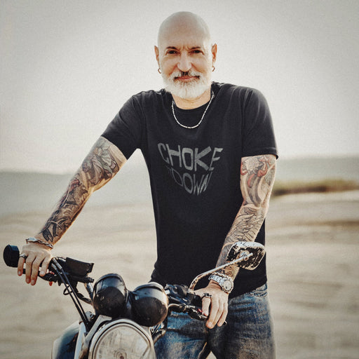 PRODUCT IMAGE: Man with tattoos and a beard standing next to a motorcycle in a desert setting wearing the Choke It Down tee | for Horny Gay Guys who appreciate oral play with varying degrees of restrained forcefulness 
