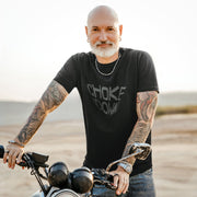 PRODUCT IMAGE: Man with tattoos and a beard standing next to a motorcycle in a desert setting wearing the Choke It Down tee | for Horny Gay Guys who appreciate oral play with varying degrees of restrained forcefulness 