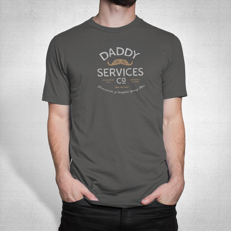 A man wearing a dark gray crewneck t-shirt with a vintage-style graphic that reads 'DADDY SERVICES CO' with a small mustache illustration, set against a light gray textured background.