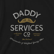 A close-up of a black t-shirt featuring a vintage-style graphic print that reads 'DADDY SERVICES CO' in distressed white and gold lettering, complete with a gold mustache illustration.