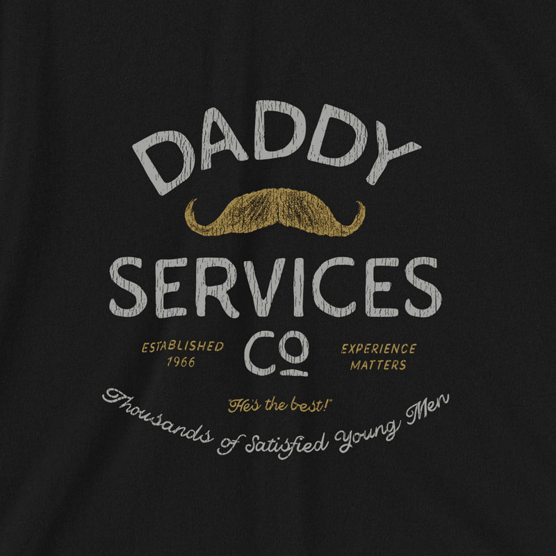 A close-up of a black t-shirt featuring a vintage-style graphic print that reads 'DADDY SERVICES CO' in distressed white and gold lettering, complete with a gold mustache illustration.