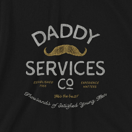 A close-up of a black t-shirt featuring a vintage-style graphic print that reads 'DADDY SERVICES CO' in distressed white and gold lettering, complete with a gold mustache illustration.