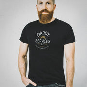 A man with a full beard wearing a black crewneck t-shirt with a white graphic that reads 'DADDY SERVICES CO' and a small mustache illustration, paired with dark blue jeans, standing against a light gray textured wall.