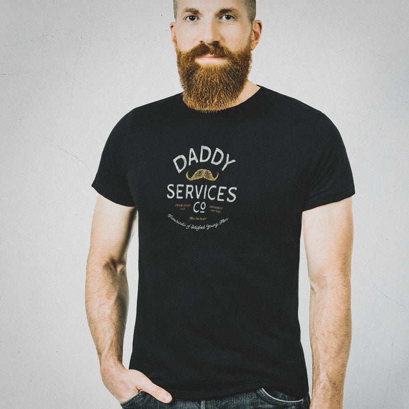 A man with a full beard wearing a black crewneck t-shirt with a white graphic that reads 'DADDY SERVICES CO' and a small mustache illustration, paired with dark blue jeans, standing against a light gray textured wall.