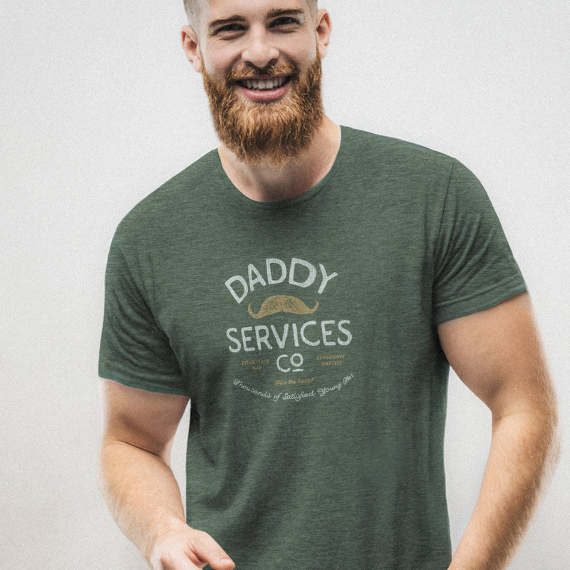 A smiling bearded man wearing a dark heather green t-shirt with a vintage-style graphic that reads 'DADDY SERVICES CO' in white and pale orange letters, posed against a neutral light gray wall.