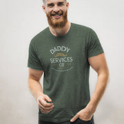 A smiling bearded man wearing a dark green graphic t-shirt with 'Daddy Services Co' and a mustache design printed on the chest, posed against a neutral light gray background.