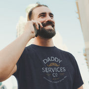 A smiling bearded man wearing a navy blue graphic t-shirt that reads 'DADDY SERVICES Co' with a mustache illustration, holding a phone to his ear outdoors.