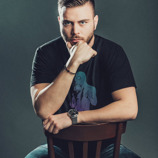 Man sitting on a chair wearing a black t-shirt with a graphic design, posing against a dark background; the image features a bold, teal & purple halftone treatment of a photo by Sergey Vinogradov depicting a partial male nude wearing a gas mask. The design, evocative of BDSM and kink culture