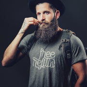 Man with a beard wearing a gray, Graphite Heather t-shirt with 'Free Refills' typographic design printed on front and a backpack; on a dark background