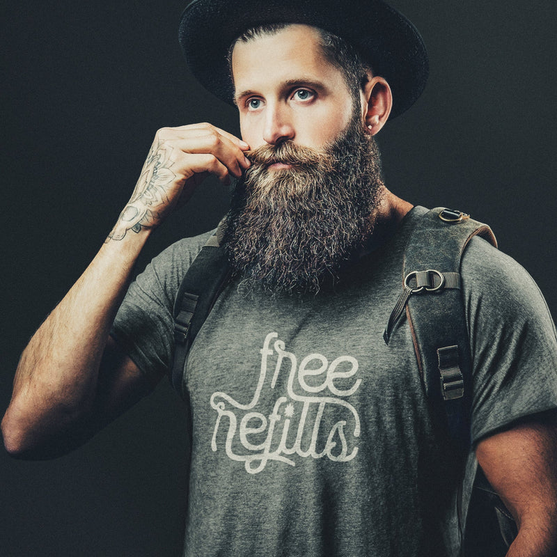 Man with a beard wearing a gray, Graphite Heather t-shirt with 'Free Refills' typographic design printed on front and a backpack; on a dark background