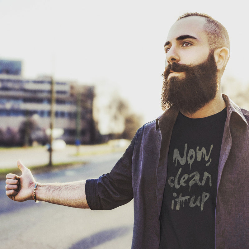 Bearded, male model attempting to hitch hike, wearing a black t-shirt with "Now, clean it up" printed on the chest; the  milky white letterforms drip, lending to an appropriate interpretation