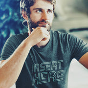 PRODUCT IMAGE: 'Insert Here' T-shirt | Suggestively raunchy tee for horny gay bottom guys | Handsome male model, with hand on chin, looking up towards top right corner wears Dark Heather tee with a white distressed typographic design is printed on it, in the chest area 