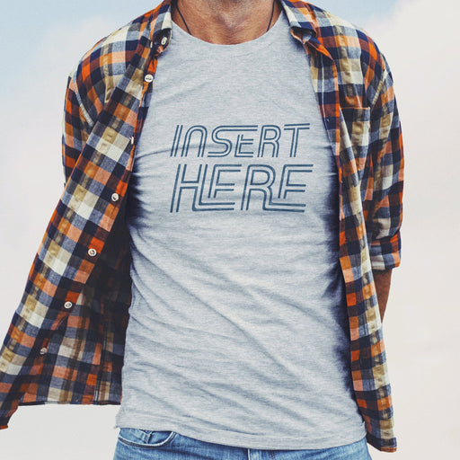 PRODUCT IMAGE: Model wears a Light Gray Heather t-shirt, with a long-sleeve plaid flanel shirt over it; unbuttoned, the top layer reveals the typographic design 'Insert Here' printed on shirt in muted dark navy letters