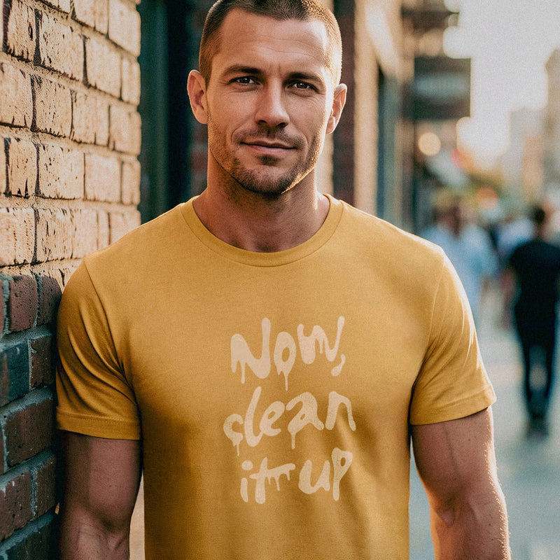Muscular model leans against brick wall on a city street wearing a mustard t-shirt with "Now, clean it up" printed on the chest; the  milky white letterforms drip, lending to an appropriate interpretation