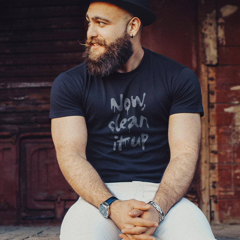 Bearded, muscular model sits on ledge, wearing a black hat and a navy t-shirt with "Now, clean it up" printed on the chest; the  milky white letterforms drip, lending to an appropriate interpretation