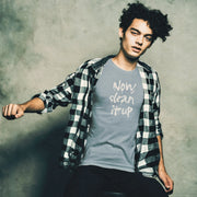 Young, thin male model sits on a sits on a stool, wearing a steel blue t-shirt with "Now, clean it up" printed on the chest; the  milky white letterforms drip, lending to an appropriate interpretation