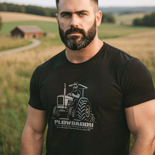 A hunky muscle daddy wears black t-shirt w/ vintage-style graphic printed on front: with bold lines, thick type that reads "Plow Daddy", and a bearded boss behind the wheel. Farmland provides the backdrop.