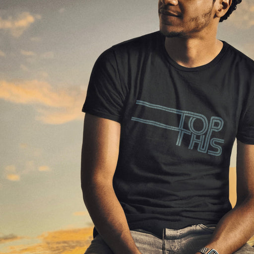 Slightly scruffy hipster male model wears 'Top This' t-shirt; black tee with a light blue, horizontal typographic design across his chest; situated against a late afternoon sky in the background