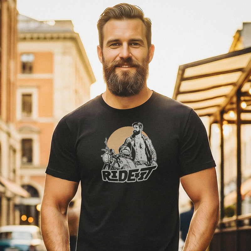 A sexy bearded model poses on a city sidewalk wearing a black t-shirt with a graphic featuring a rugged, bearded biker in a distressed vintage print style with the design's namesake, "Ride It."