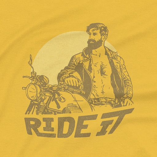 A close-up detail of the graphic printed on a mustard tee featuring a rugged, bearded biker in a distressed vintage print style with the design's namesake, "Ride It."
