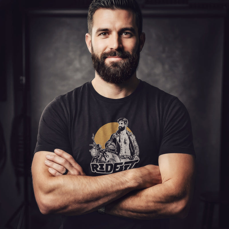 A sexy bearded model poses in a studio wearing a dark gray t-shirt with a graphic featuring a rugged, bearded biker in a distressed vintage print style with the design's namesake, "Ride It."