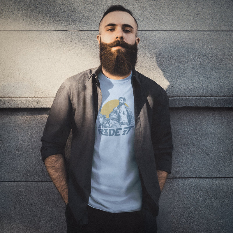 A sexy bearded model poses against a concrete wall wearing a light blue t-shirt with a graphic featuring a rugged, bearded biker in a distressed vintage print style with the design's namesake, "Ride It."