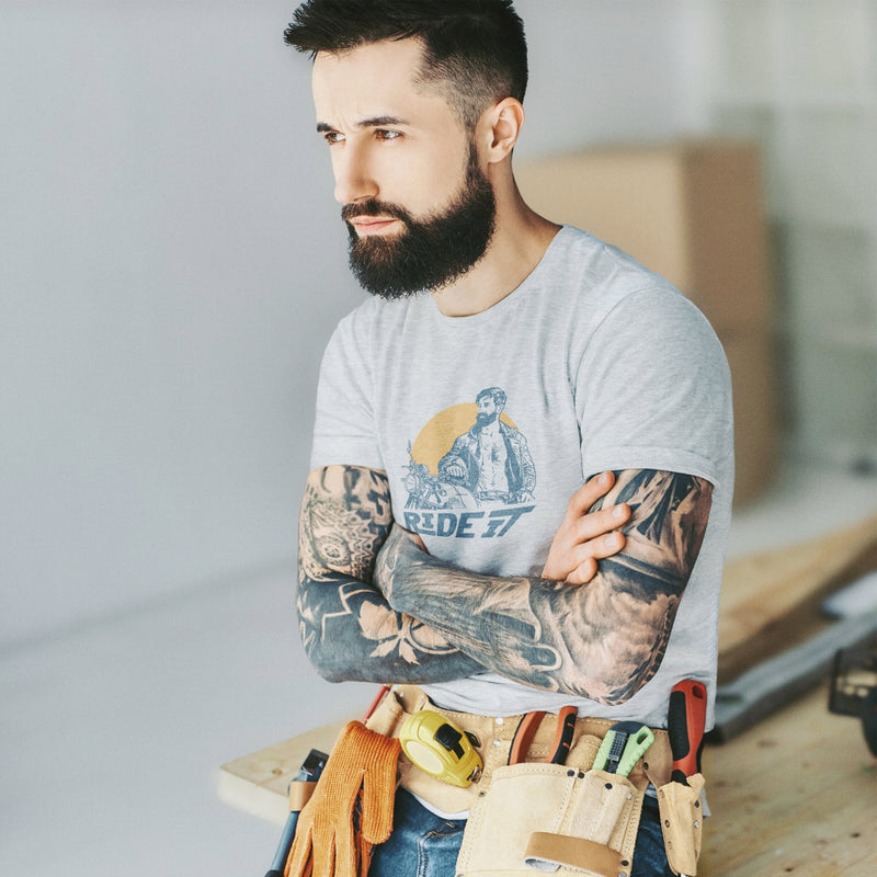 A sexy bearded model poses on the work site wearing a light gray heather t-shirt with a graphic featuring a rugged, bearded biker in a distressed vintage print style with the design's namesake, "Ride It."