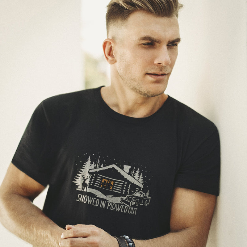 Men’s graphic t-shirt showing a snowy log cabin and plow truck, featuring the phrase “Snowed In, Plowed Out,” a subtle innuendo design for gay men; Model wears black tee.
