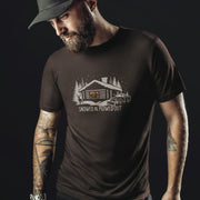 Men’s graphic t-shirt showing a snowy log cabin and plow truck, featuring the phrase “Snowed In, Plowed Out,” a subtle innuendo design for gay men. Bearded male model wears brown tee.