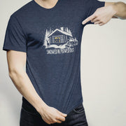 Men’s graphic t-shirt showing a snowy log cabin and plow truck, featuring the phrase “Snowed In, Plowed Out,” a subtle innuendo design for gay men. Bearded male model wears heather navy tee.